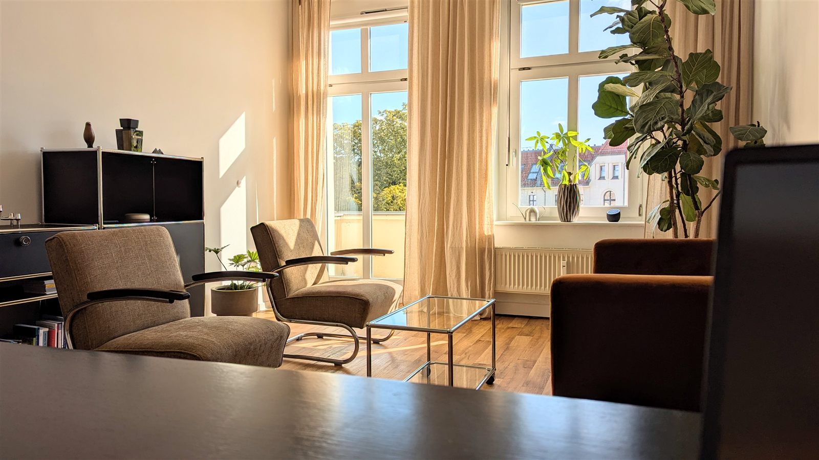 Blick vom Schreibtisch in den Praxisraum: zwei Sessel, ein Glastisch und hohe Fenster im Sonnenlicht